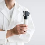 male board-certified dermatologist in a white lab coat holding a dermatoscope